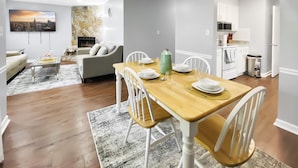 A modern living room with a fireplace, a dining area with a wooden table set for four, and a kitchen with white cabinets. - NEW! Renovated Home - 10 Min to Augusta Downtown! (Evans)