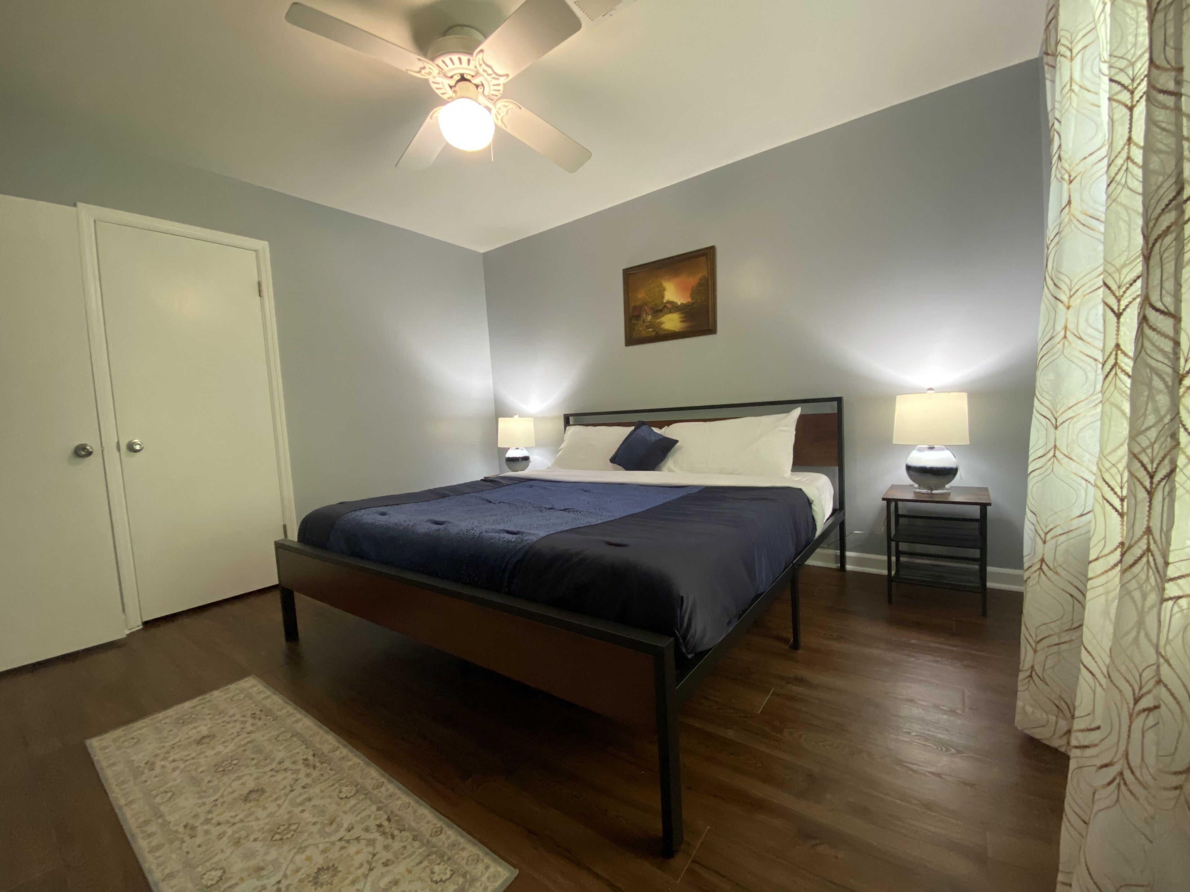 A bedroom with a bed, two bedside tables with lamps, a ceiling fan, a painting on the wall, and a patterned curtain.