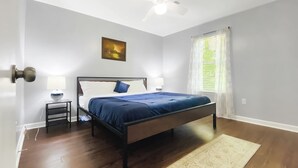 A bedroom with a bed, bedside tables, a nightstand, a painting, a window with curtains, and a wooden floor. - NEW! Renovated Home - 10 Min to Augusta Downtown! (Evans)