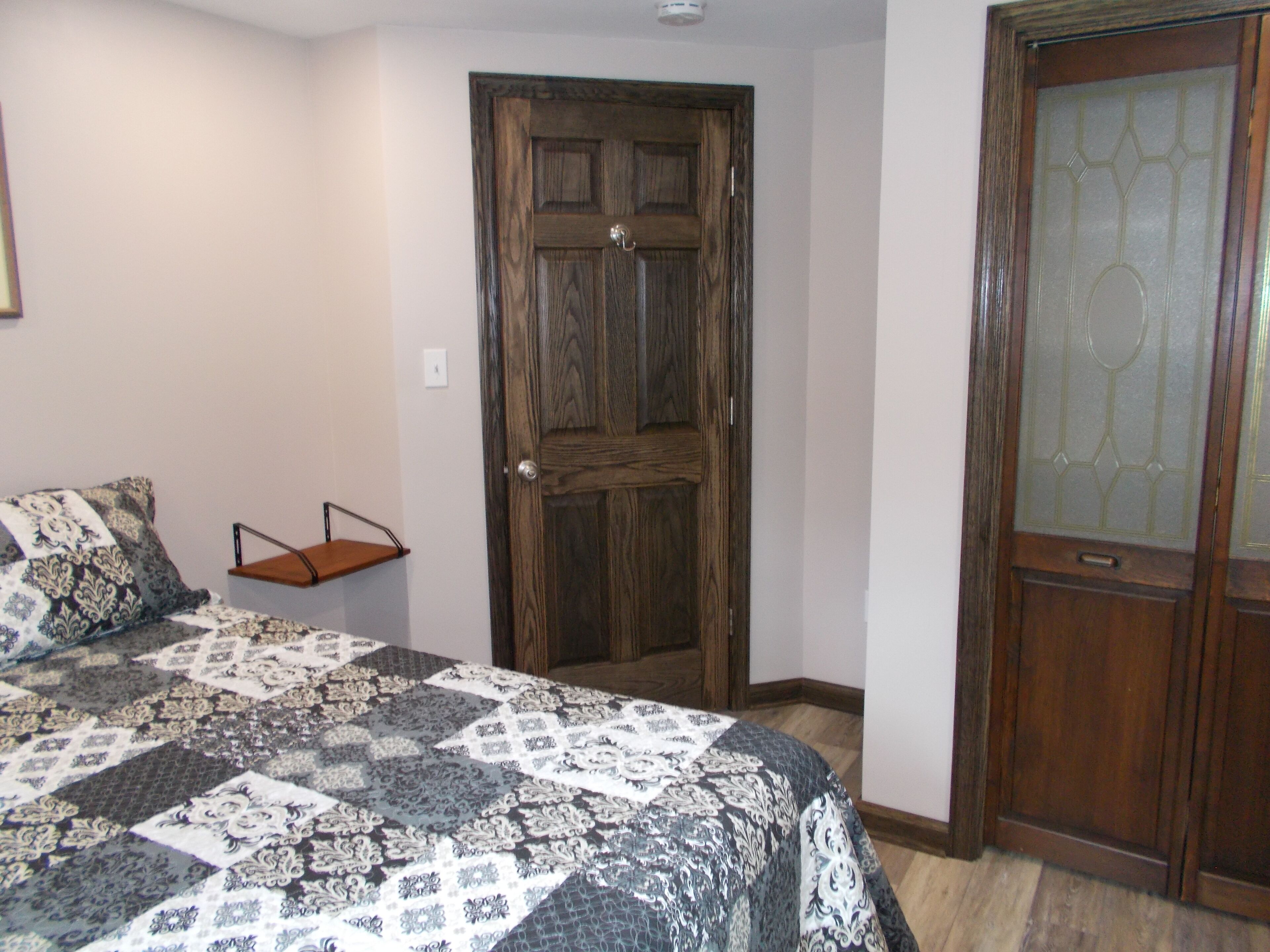 Quarto com porta de madeira, cama com colcha estampada e pequena estante de madeira.