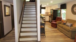 Eine Treppe führt zu einer zweiten Etage, einem Küchenbereich mit Kühlschrank, einem Esstisch und einem Sofa mit einem 'COZY' Kissen.