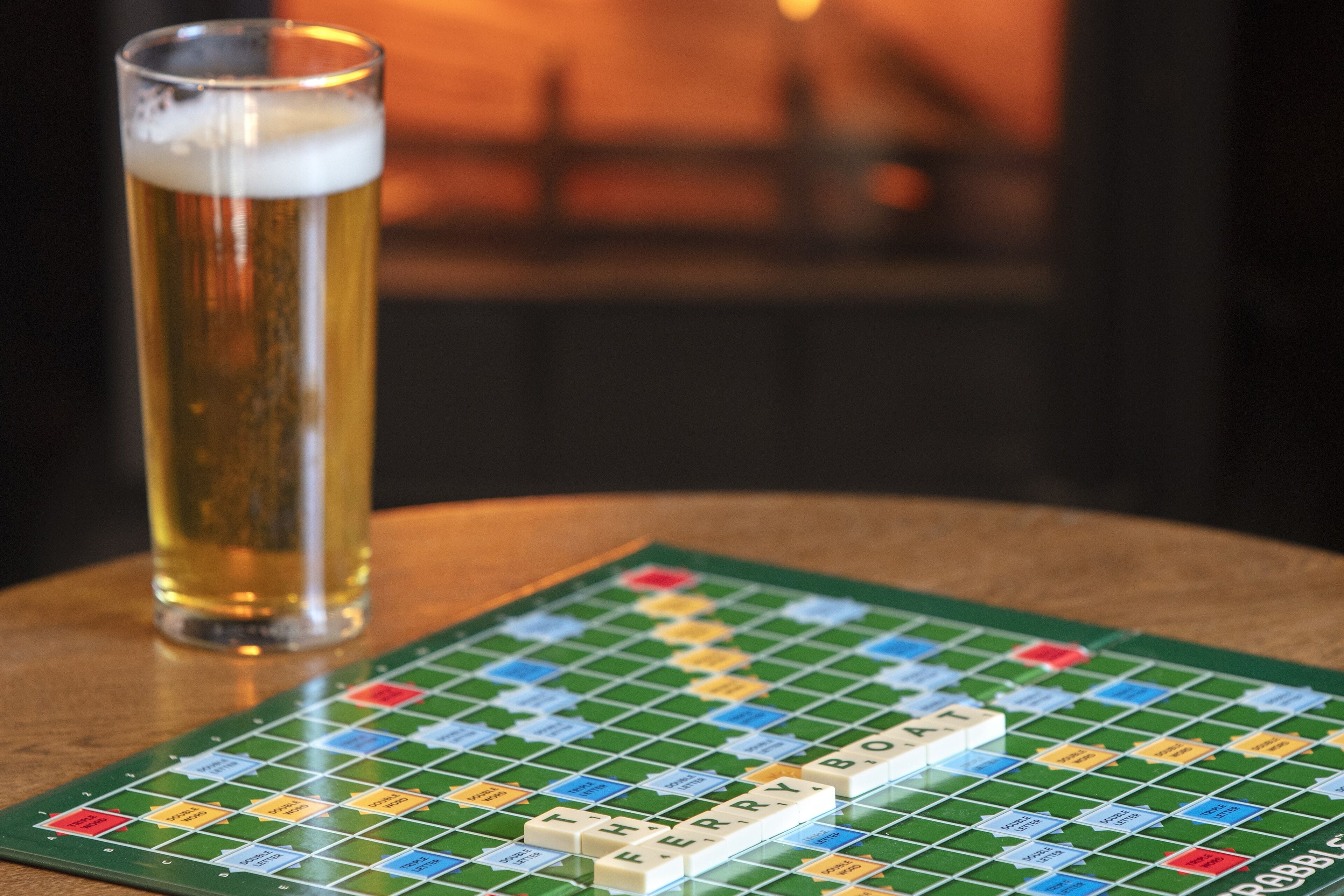 Ein Scrabble-Spielbrett mit dem Wort 'FERRYBOAT', ein Glas Bier und ein unscharfer Hintergrund, der eine gemütliche Innenraumgestaltung andeutet.