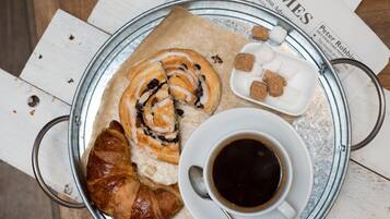 A tray with a croissant, a cinnamon roll, a cup of coffee, and a small dish of sugar cubes on a wooden table with newspapers.