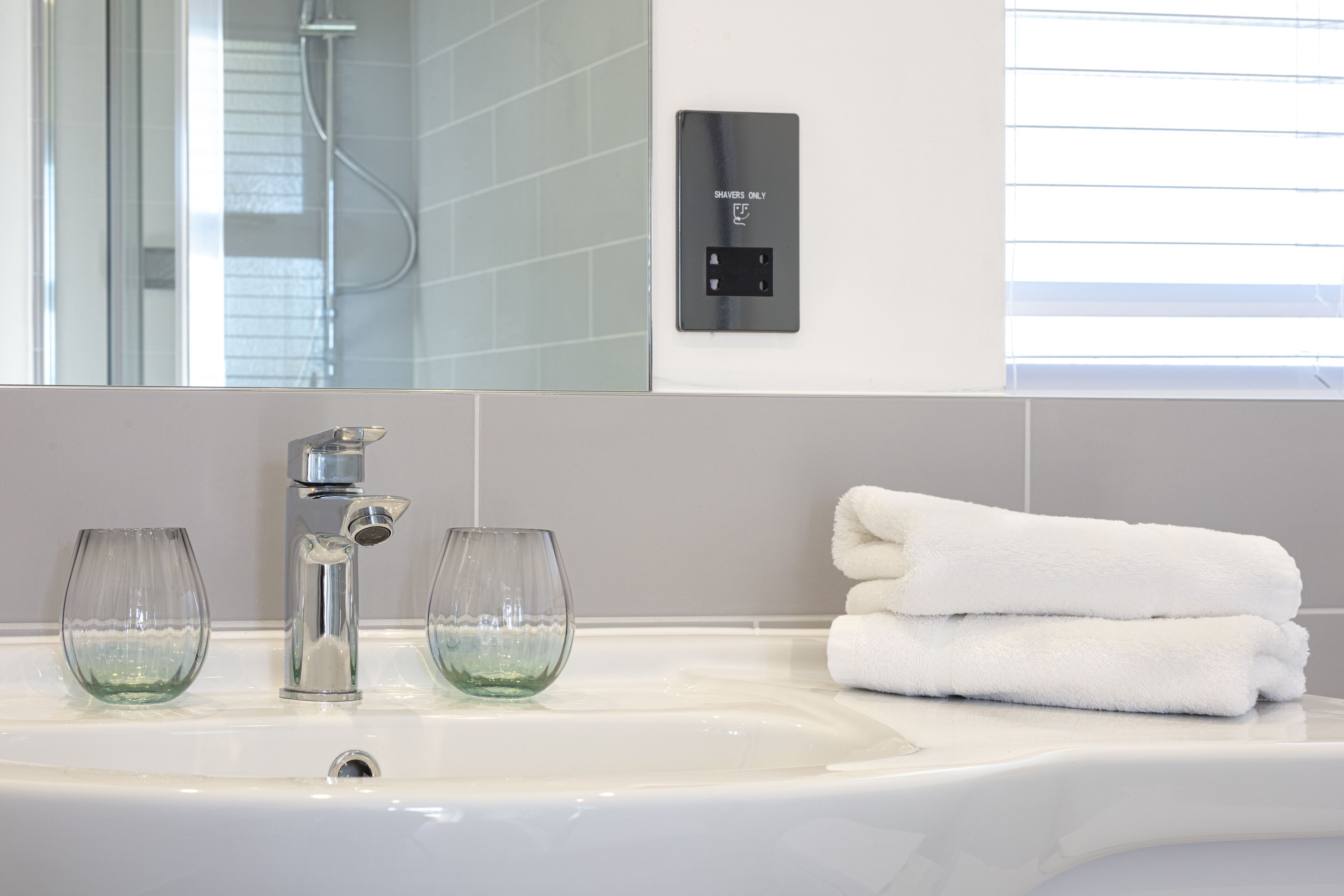 A bathroom with a white sink, two clear glasses, a towel stack, and a wall-mounted control panel.