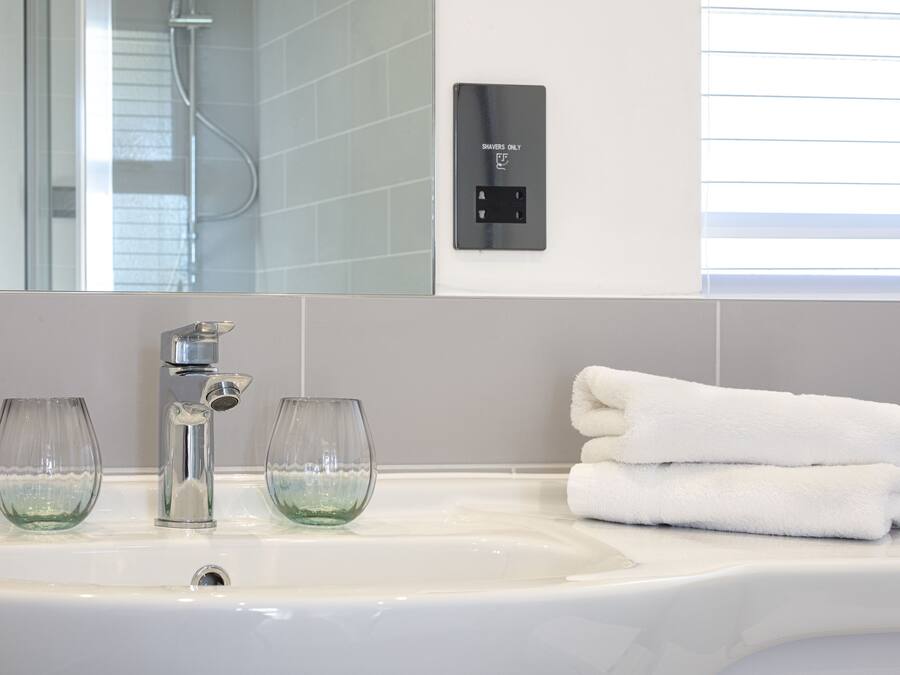 A bathroom with a white sink, two clear glasses, a towel stack, and a wall-mounted control panel.