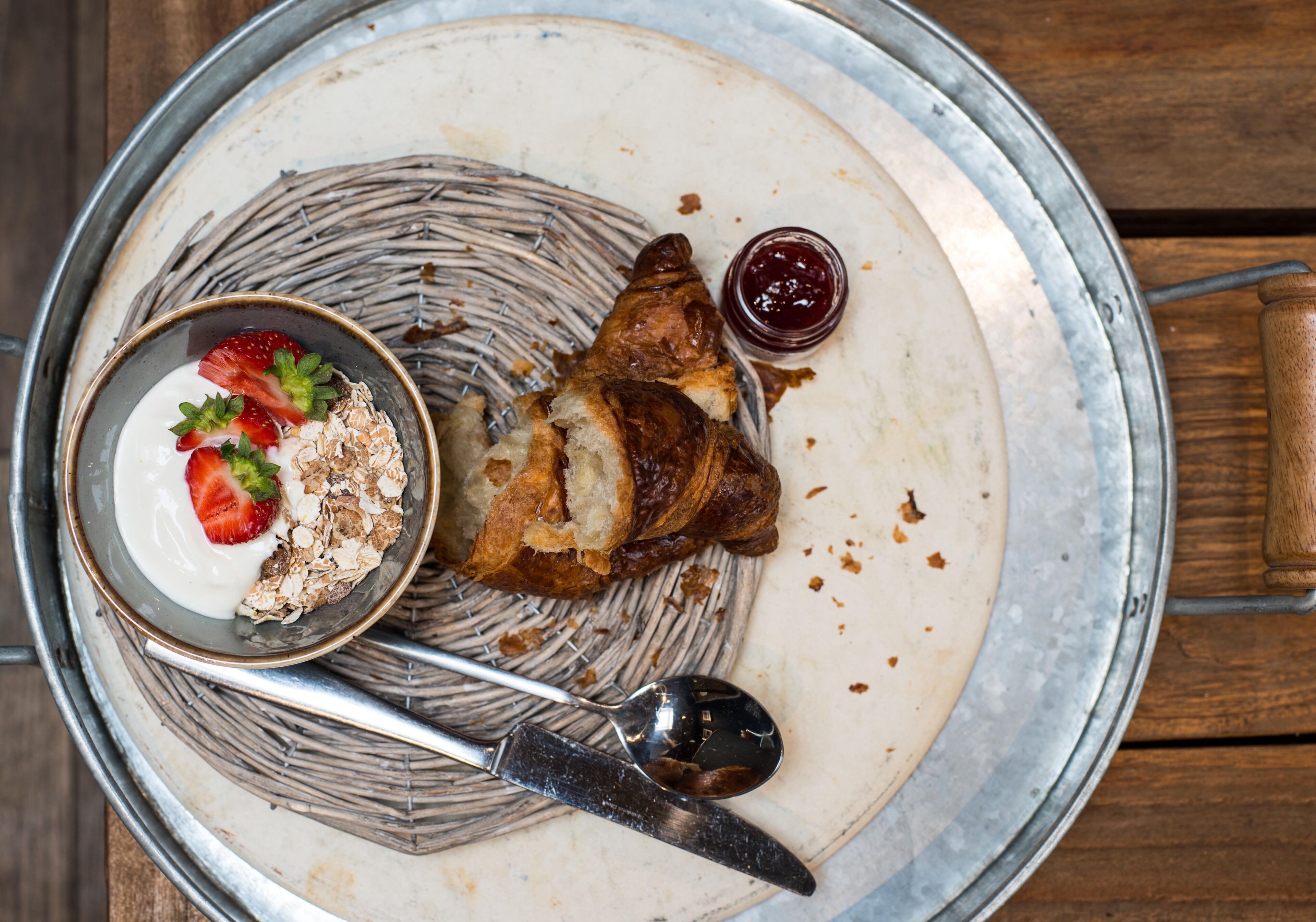 Un vassoio di metallo con un piatto contenente un croissant, una ciotola di yogurt e fragole, e un cucchiaio.