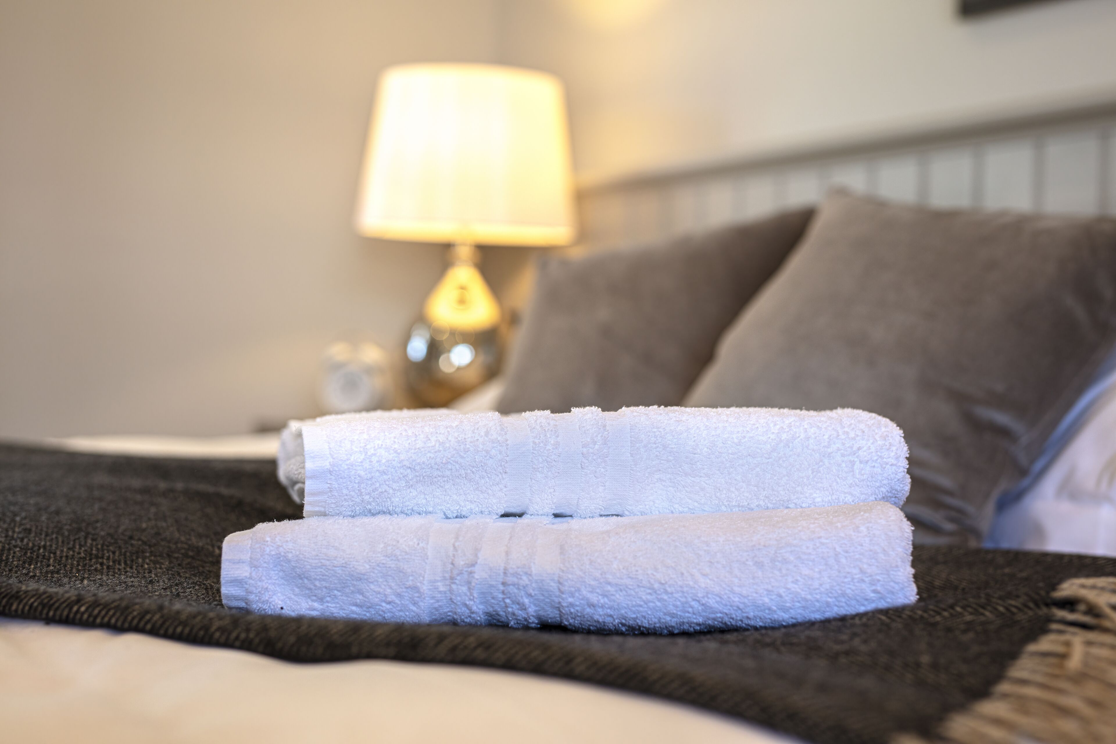 A neatly made bed with two folded white towels, a dark blanket, and a bedside lamp.