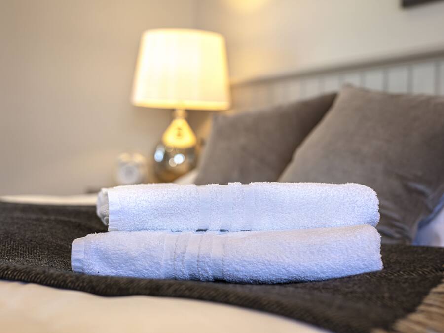 A neatly made bed with two folded white towels, a dark blanket, and a bedside lamp.