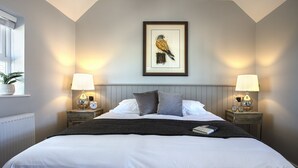A neatly made bed with white linens, two bedside tables with lamps, a framed picture of a bird on the wall, and a window with a plant. - The Ferry Boat Inn (Chelmsford)