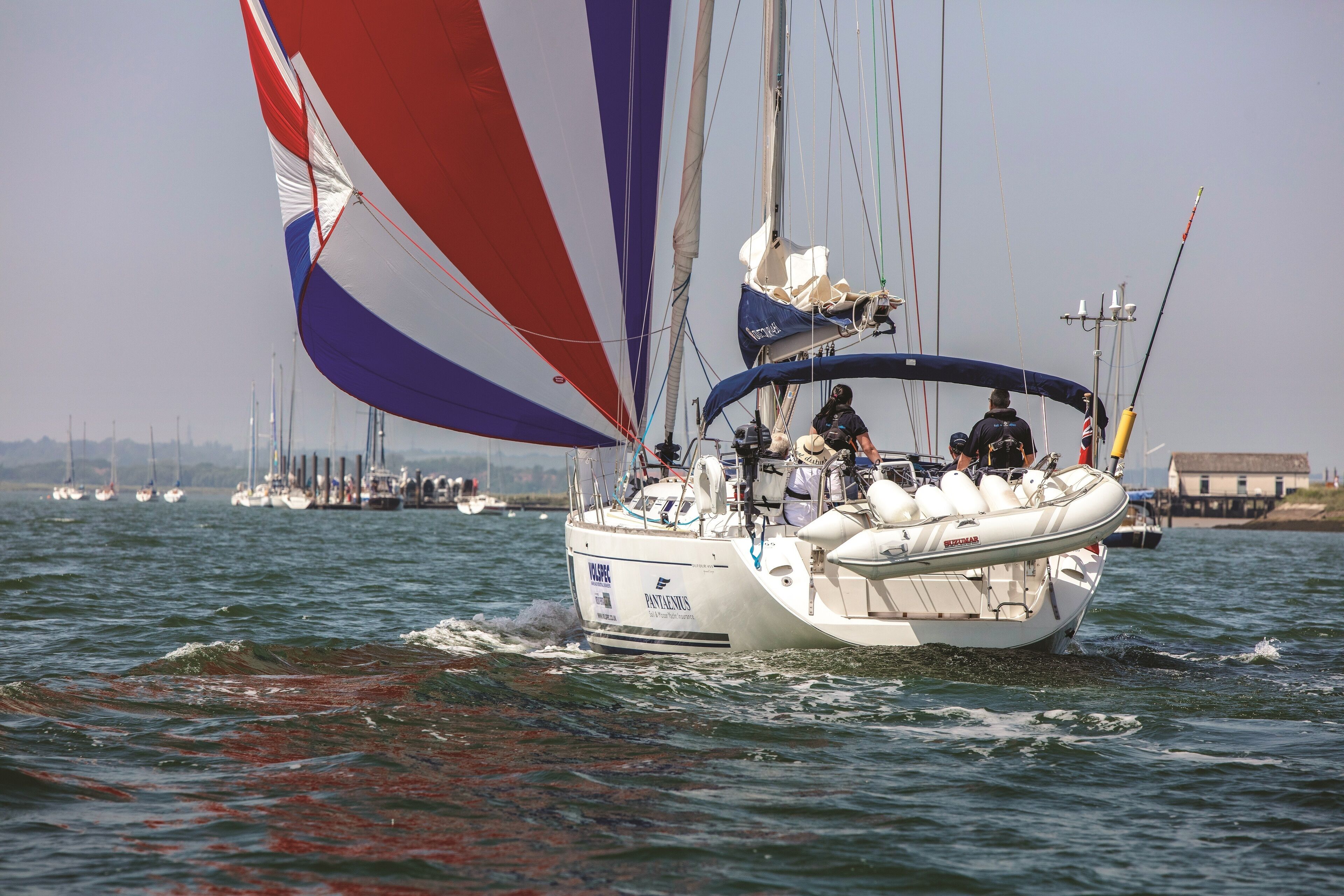 Un voilier à la coque blanche et à la voile bleue et rouge, naviguant sur une étendue d’eau, avec d’autres bateaux en arrière-plan.