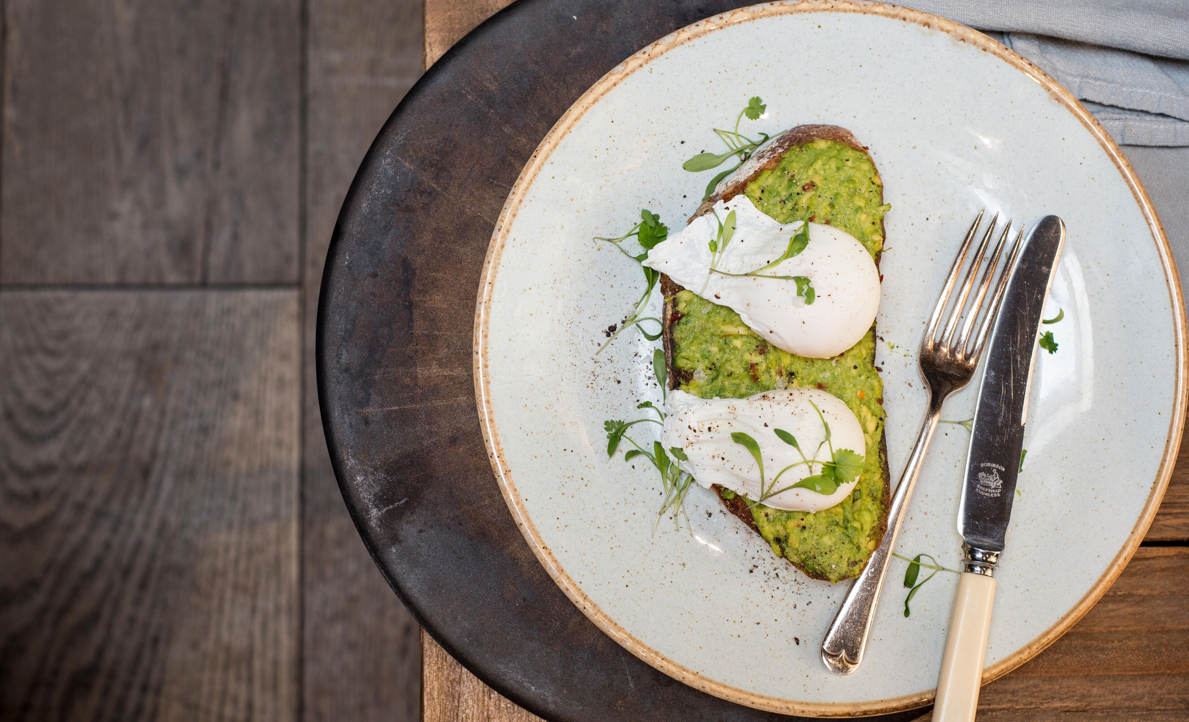 Un plato con una rebanada de tostada cubierta de guacamole y un huevo pochado, decorado con hierbas, sobre un plato de cerámica moteada.