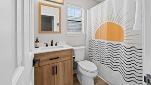 Une salle de bain avec un meuble-lavabo en bois, des toilettes blanches, un rideau de douche à motifs solaires et une fenêtre avec des stores.