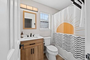 Ein Badezimmer mit einem Holzbadschrank, einer weißen Toilette, einem Sonnendekor-Duschvorhang und einem Fenster mit Jalousien.