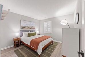 Un dormitorio con cama, mesitas de noche, lámpara, ventana con contraventanas y un cuadro colgado en la pared.