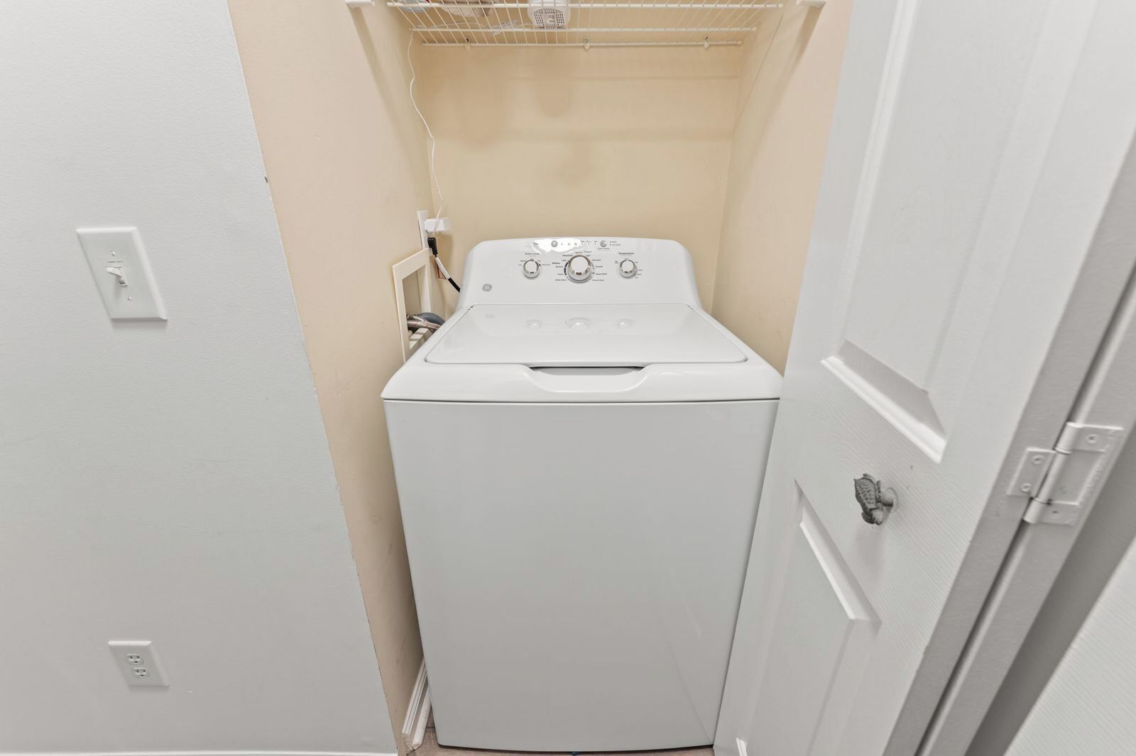 A compact laundry room with a front-loading washing machine, a power outlet, and a door.