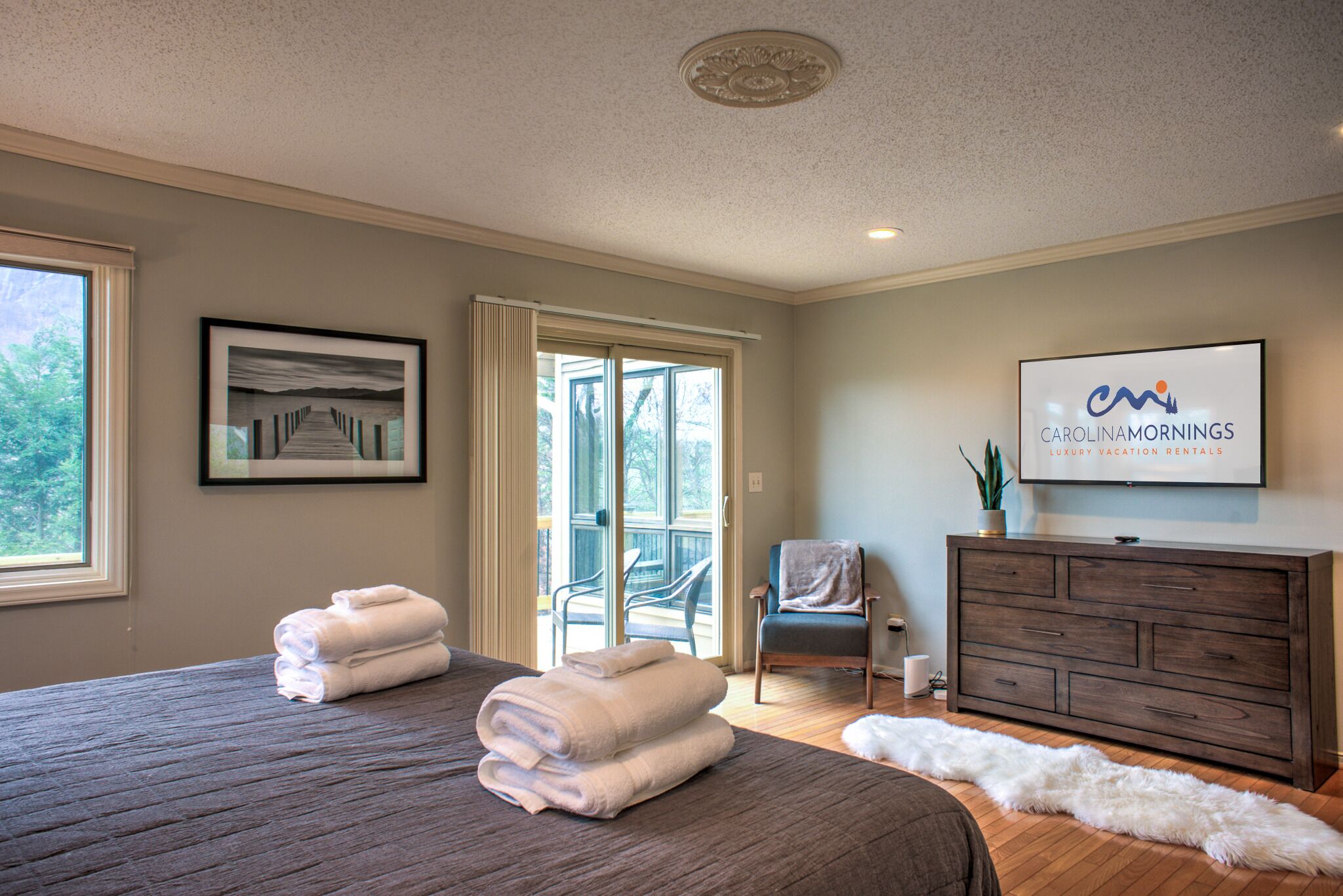 Ein geräumiges Schlafzimmer mit zwei großen Betten, einer Schiebetür zu einem Balkon, einem Fernseher mit der Anzeige 'Carolinamornings Luxury Vacation Rentals' und einem gerahmten Bild an der Wand.