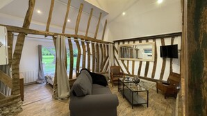 Een moderne woonkamer met zichtbare houten balken, een groot raam, een grijze bank, een houten salontafel en een flatscreen televisie die aan de muur is bevestigd.