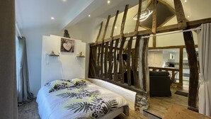 Een slaapkamer met een bed, houten balken, een stenen muur en uitzicht op de keuken.