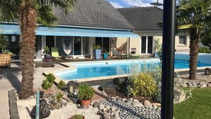 A poolside area with a pool, palm trees, and a house with a blue awning. - Escale En Correze (Sadroc)