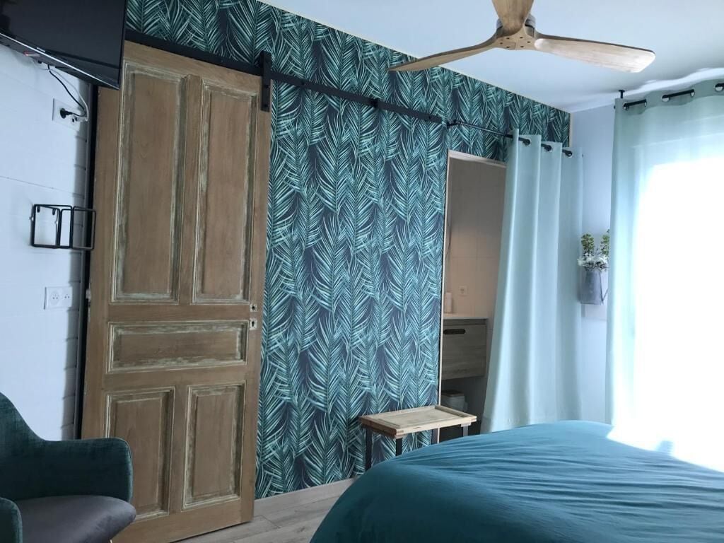A bedroom with a bed, a wooden wardrobe, a ceiling fan, a mirror, and a window with curtains.