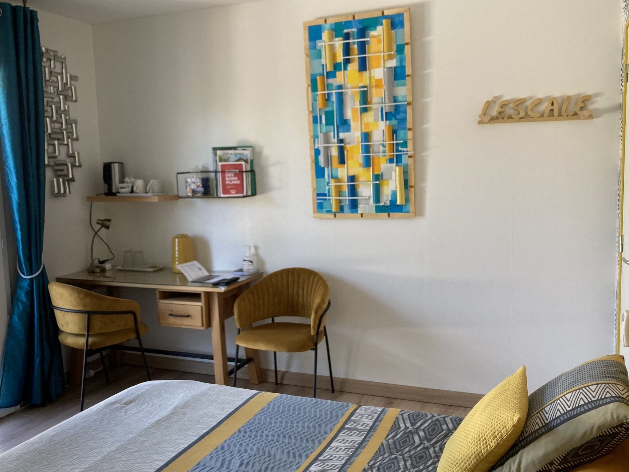 A hotel room with a bed, a desk, two chairs, a wall art piece, and a shelf with items.