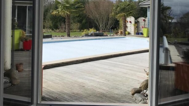 A view through a glass door of a swimming pool area with a wooden deck, palm trees, and a clear sky. - Escale En Correze (Sadroc)