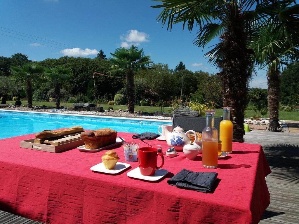 Et spisebord med morgenmadsprodukter ved en pool, omgivet af palmer og en klar himmel.