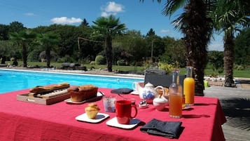 Et spisebord med morgenmadsprodukter ved en pool, omgivet af palmer og en klar himmel.