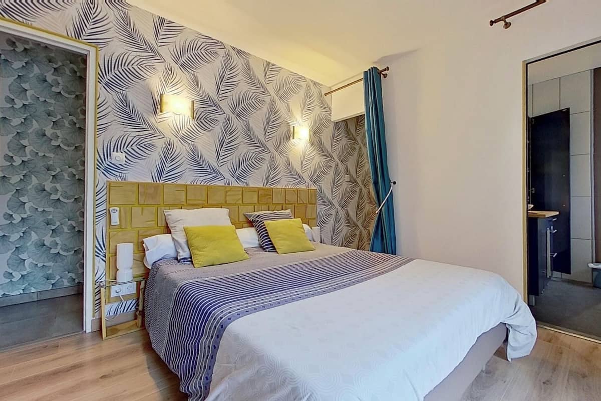 A modern bedroom with a patterned wallpaper, a wooden headboard, a bed with striped bedding, and a bathroom visible through an open door.