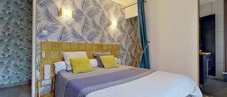 A modern bedroom with a patterned wallpaper, a wooden headboard, a bed with striped bedding, and a bathroom visible through an open door.