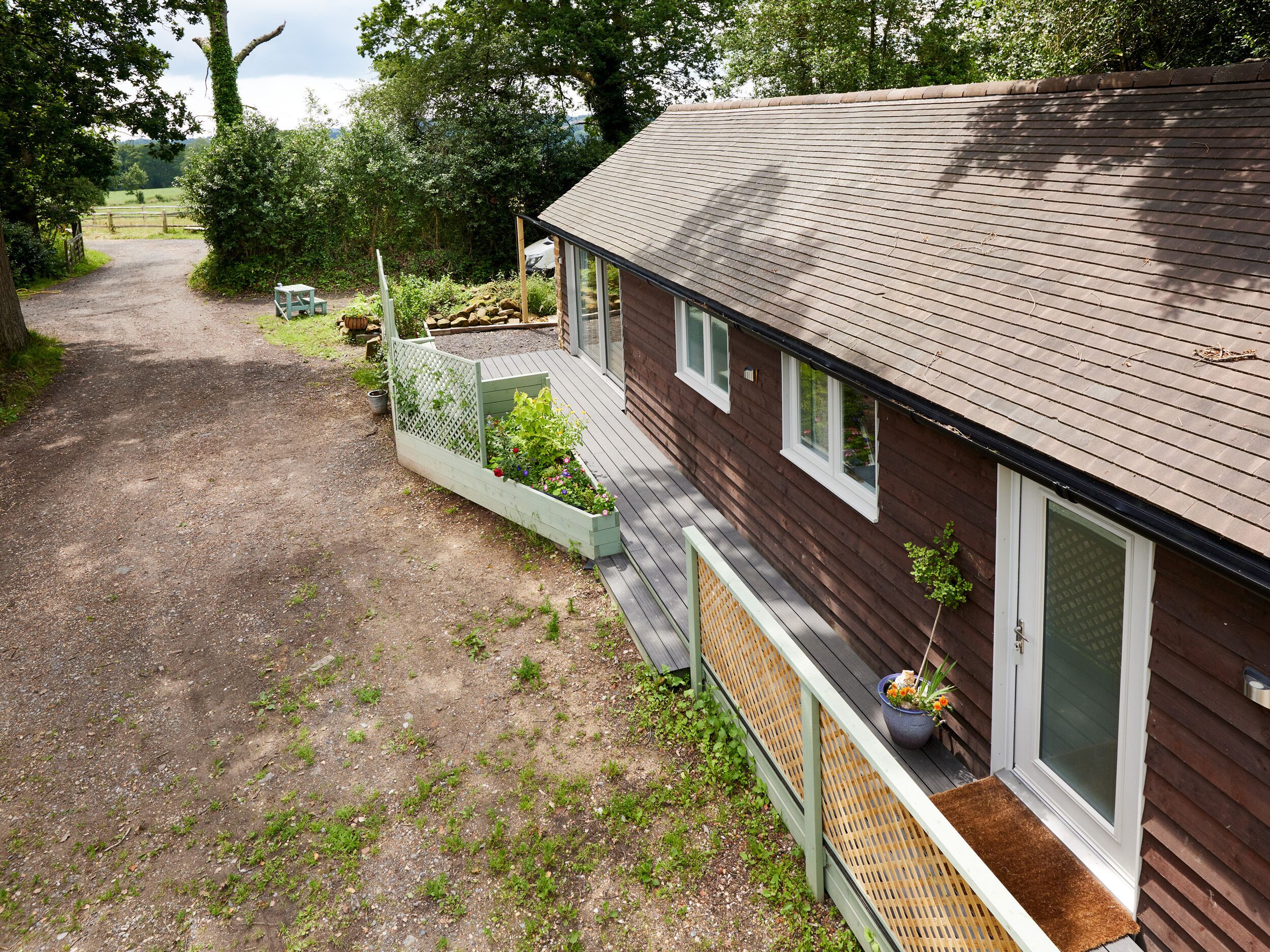 A wooden house with a gravel driveway, a wooden deck, and a garden area with plants and flowers.