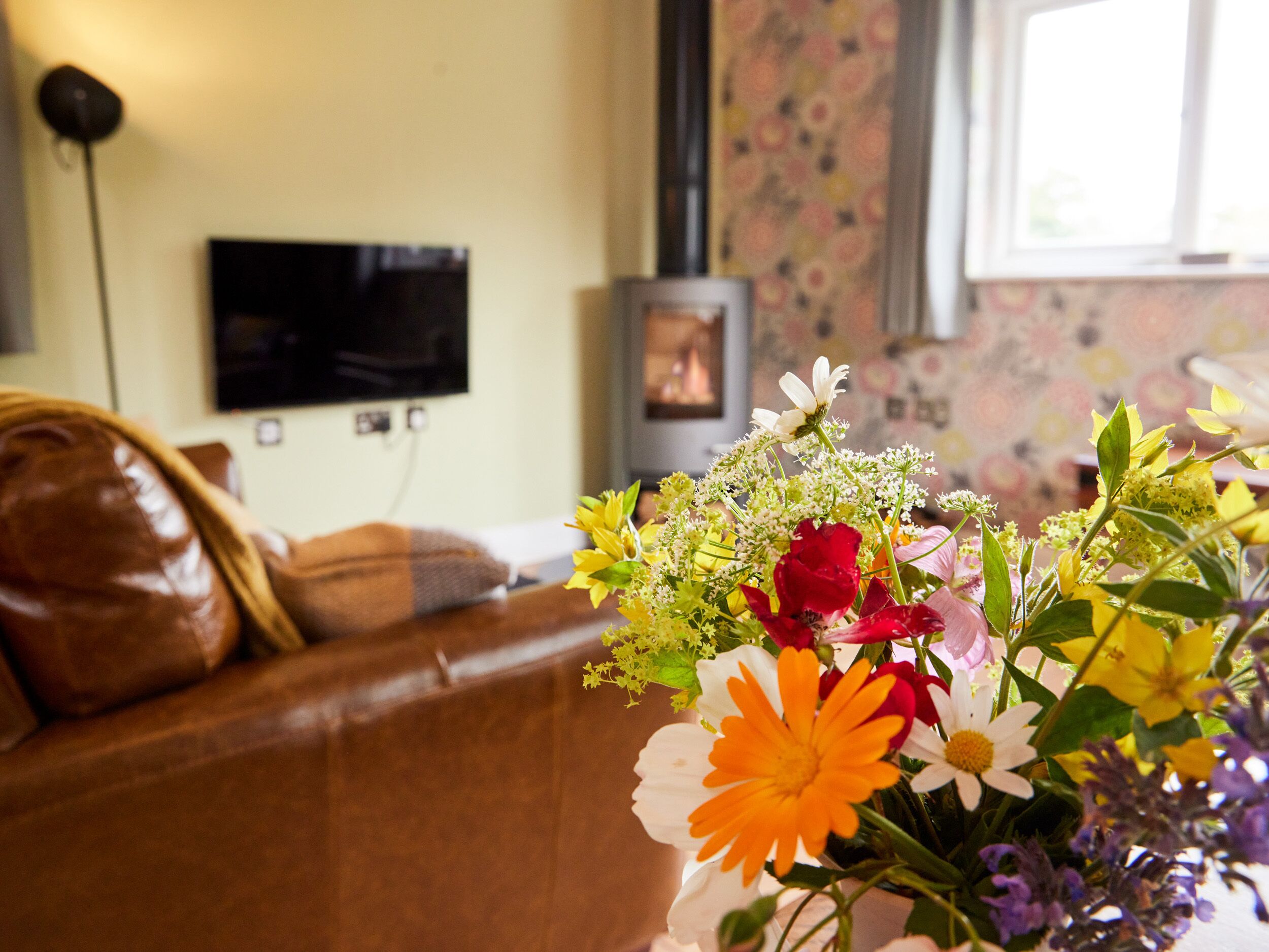 A living room with a brown leather sofa, a flat-screen TV mounted on the wall, and a floral arrangement on a table.