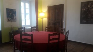 A dining area with a round table covered in a red tablecloth, surrounded by wooden chairs. There is a window with curtains, a radiator, and a wall-mounted painting.