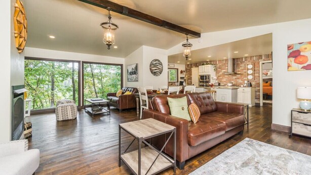 A spacious living room with a leather sofa, a coffee table, and a dining area. There is a kitchen in the background with modern appliances and a brick backsplash.