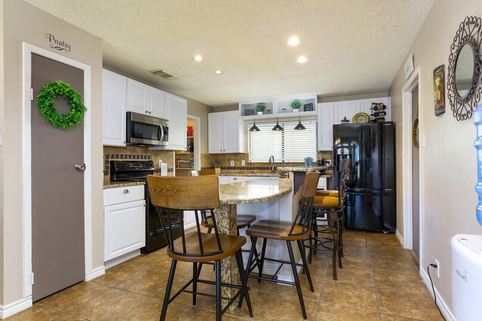 A kitchen with white cabinets, a granite countertop, a black refrigerator, and a dining area with bar stools.