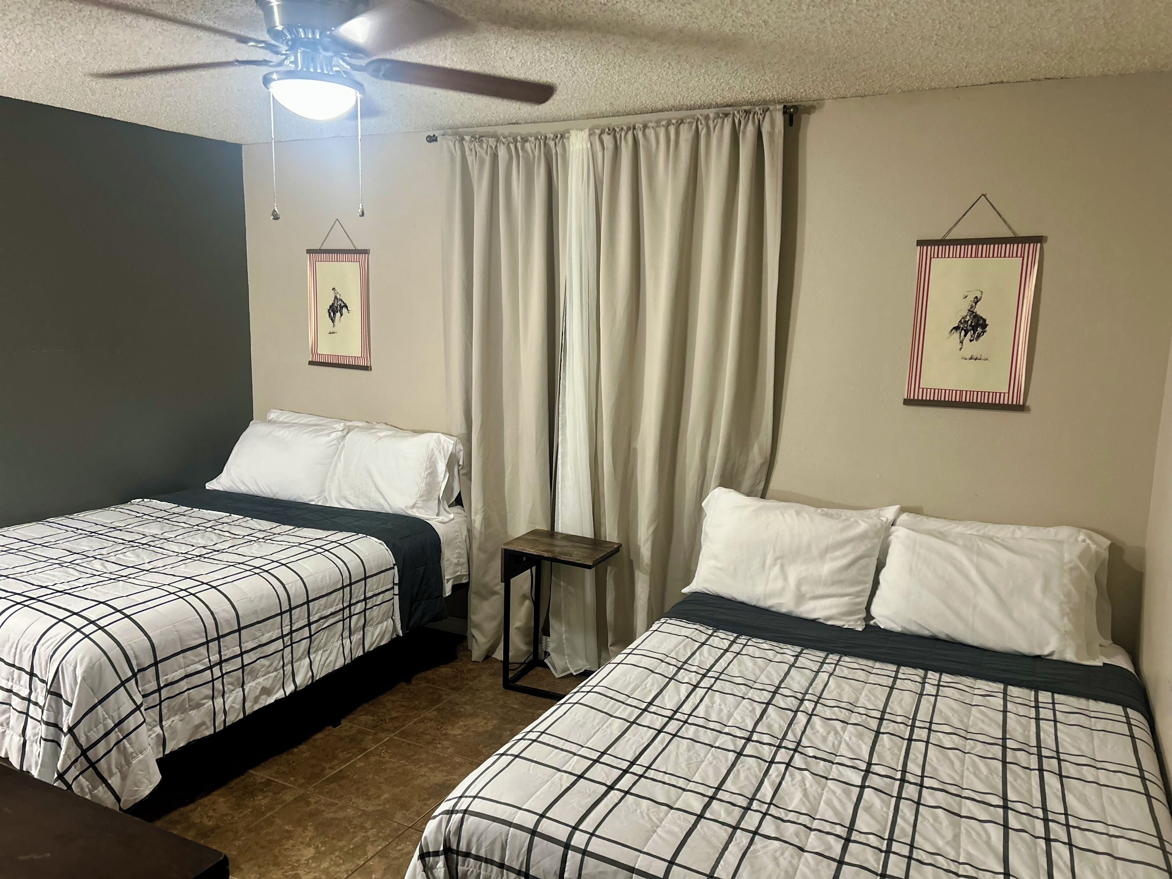 Habitación de hotel con dos camas, un ventilador de techo, una mesita y dos cuadros en las paredes.