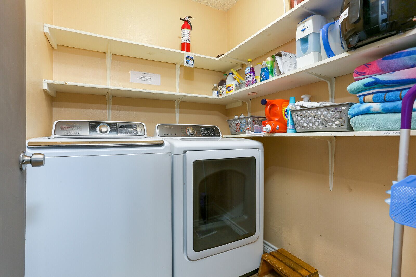 A laundry area with two washing machines, a fire extinguisher, shelves with cleaning supplies, and a microwave.