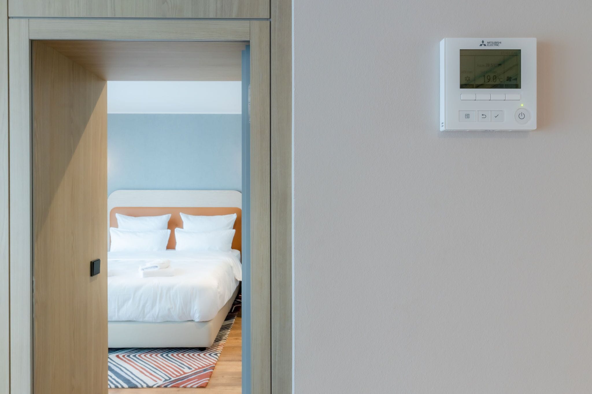 A hotel room with a bed, a wall-mounted thermostat, and a wooden door frame.