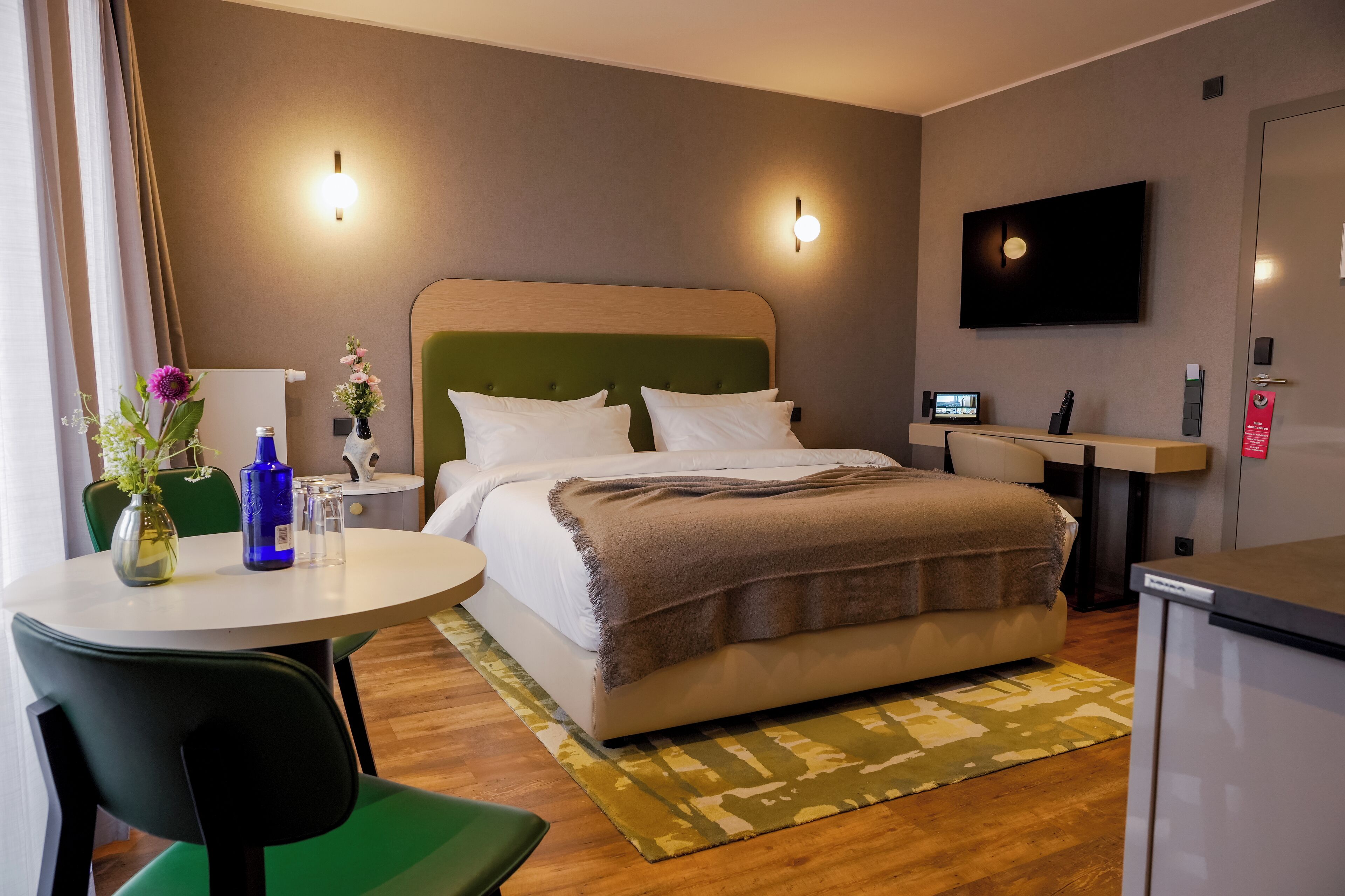 A hotel room with a bed, a desk, a chair, a TV, and a small table with a vase and flowers.