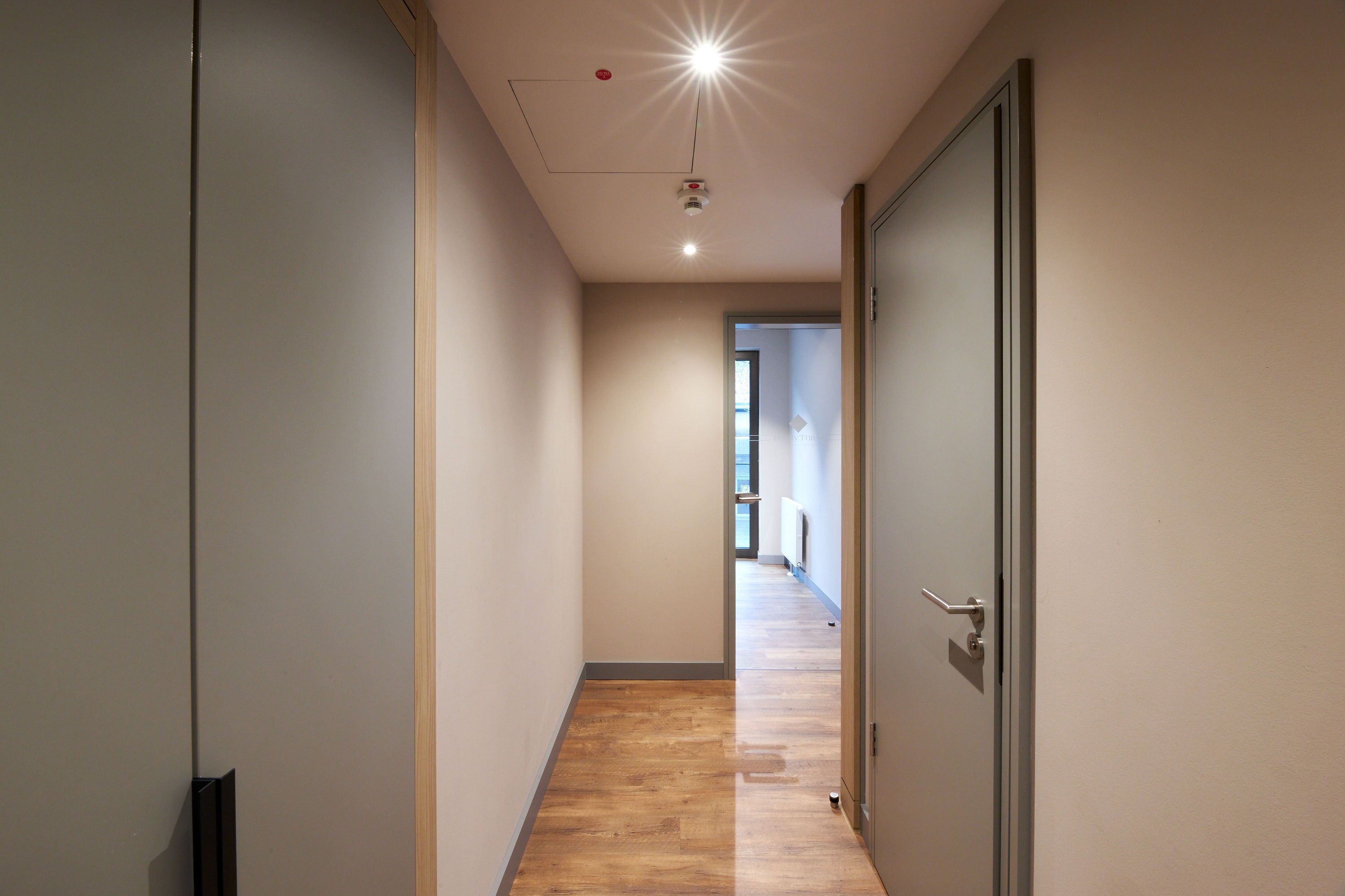 A narrow hallway with wooden flooring, a door on the right, and a ceiling light.