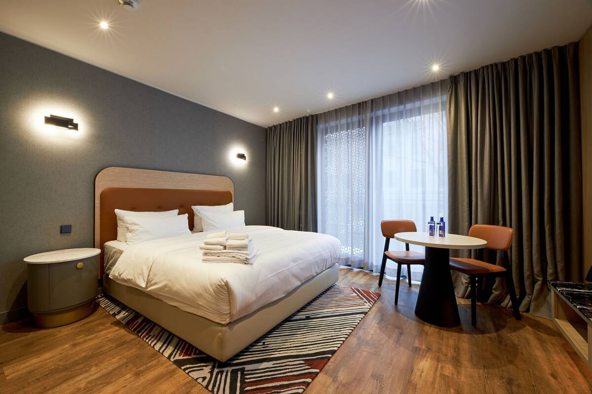 A modern hotel room with a large bed, a small table with chairs, and a view of the outdoors through a large window.