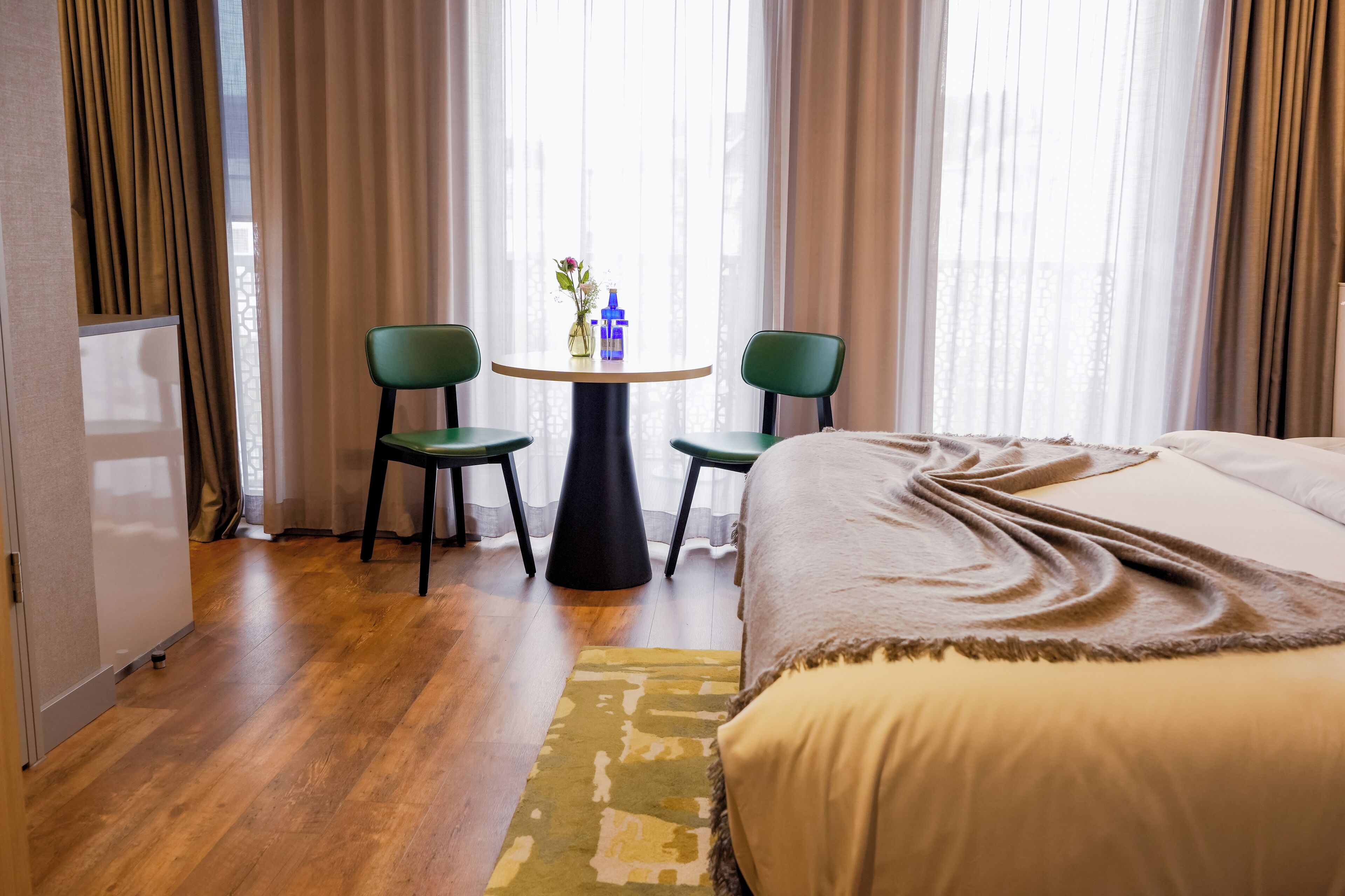 A hotel room with a bed, two chairs, a small table with a vase, and a window with curtains.