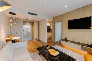 Una sala de estar moderna con un sofá, una mesa de centro y un televisor montado en la pared. Se ve un área de cocina a través de una puerta abierta.