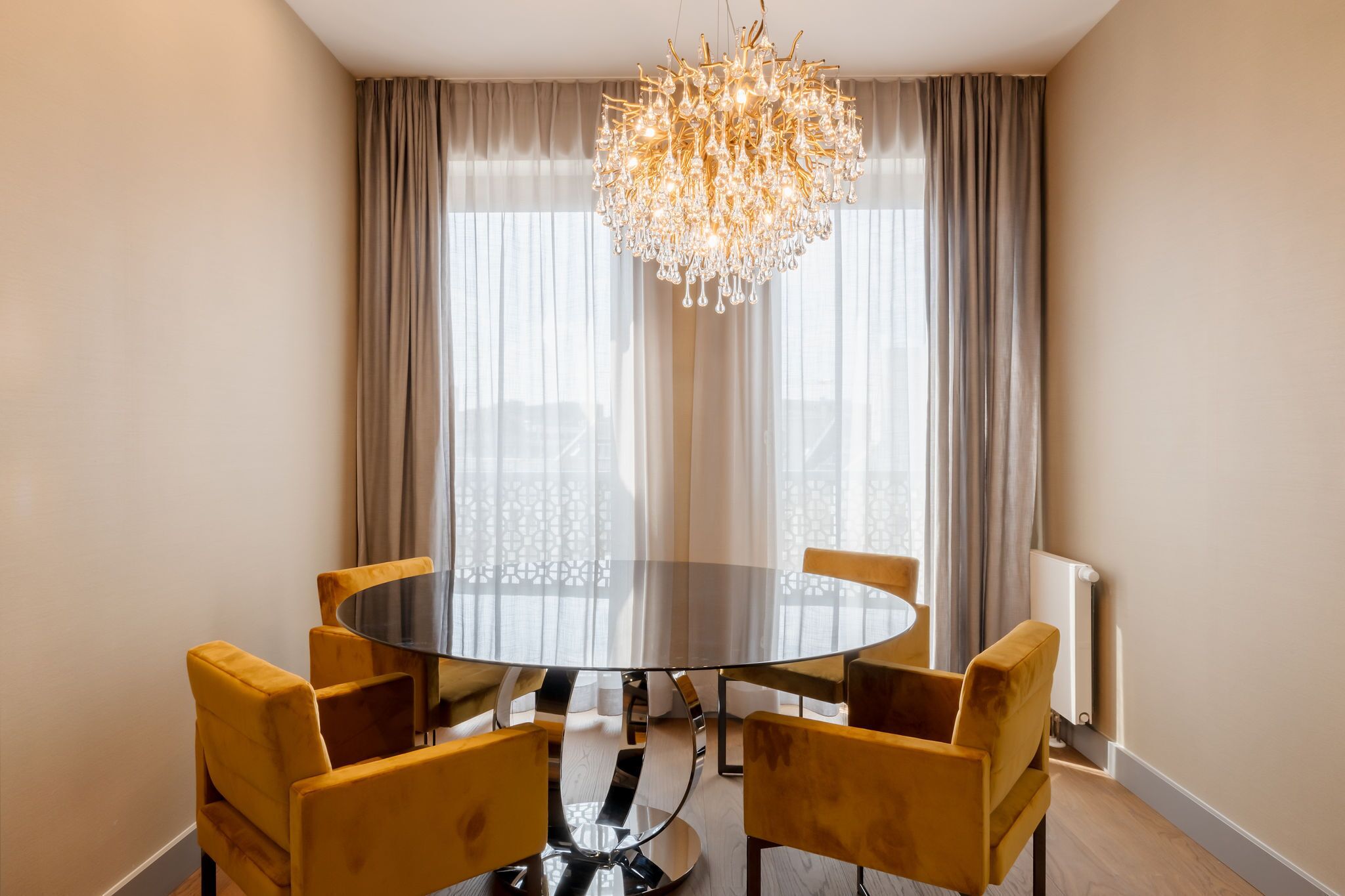 A round dining table with six chairs, a chandelier, and a view through sheer curtains.