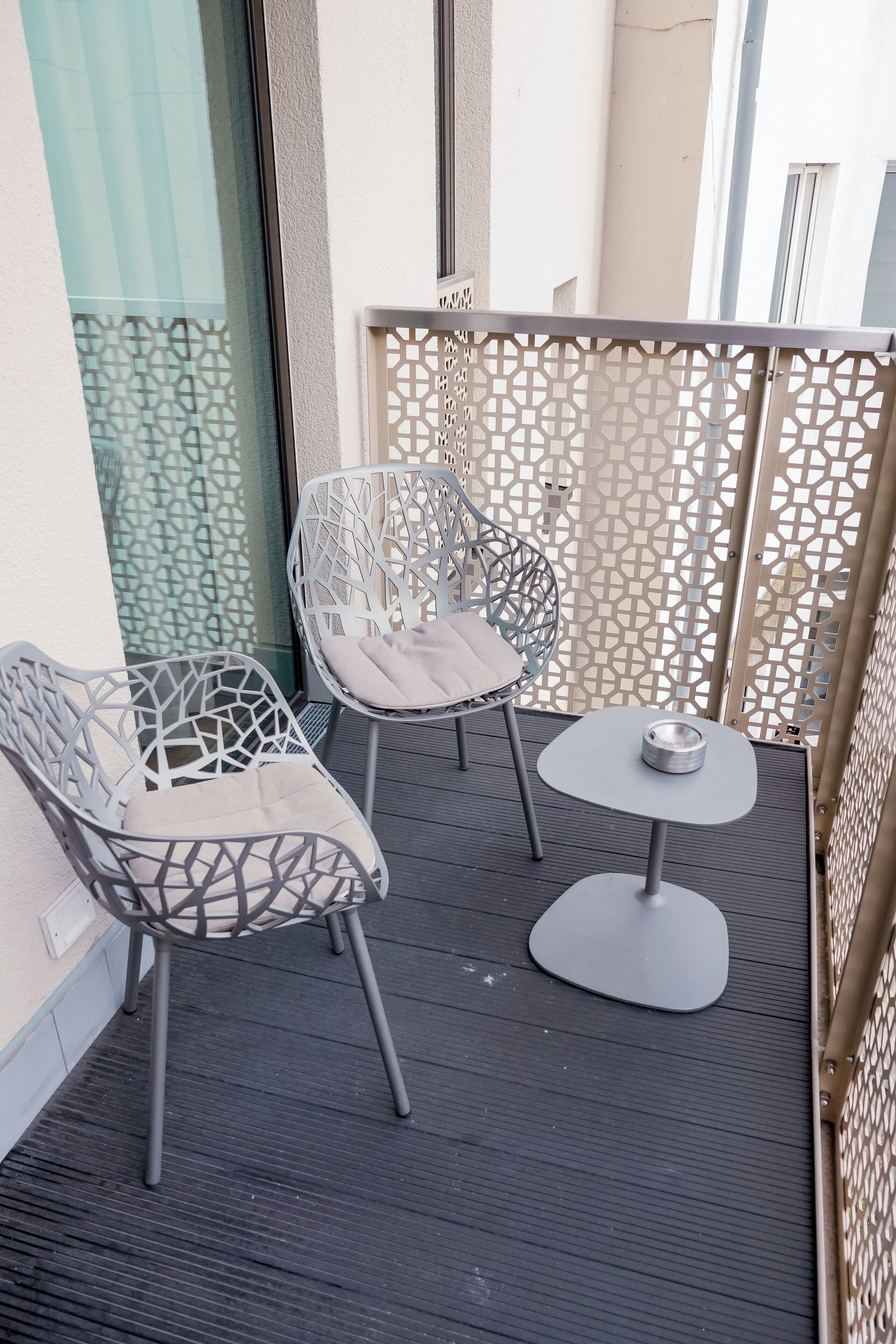 Ein Balkon mit zwei Metallstühlen, einem kleinen Tisch, einem dekorativen Metallzaun und einer Glastür.