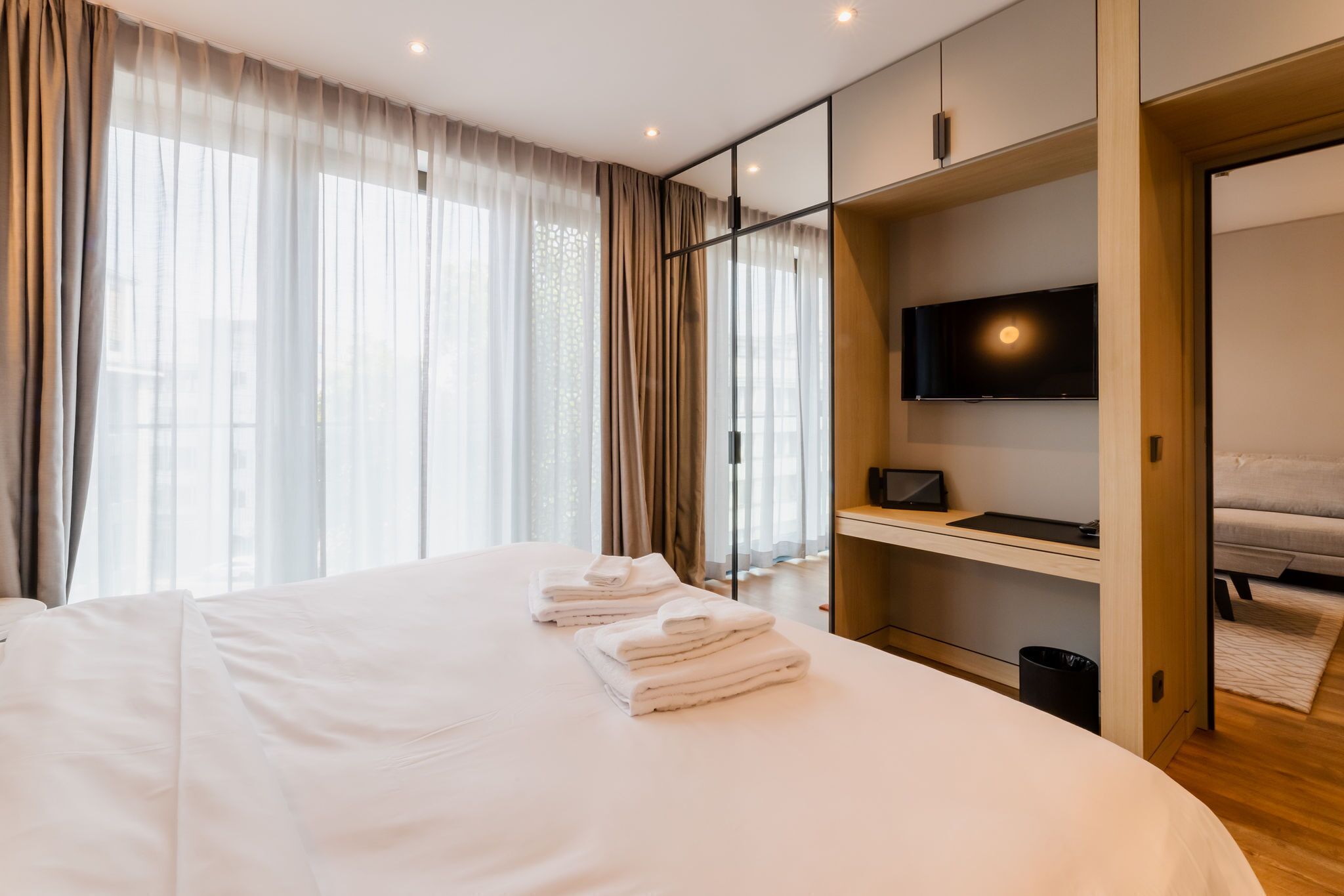 A modern hotel room with a large bed, a TV mounted on the wall, and a view of the city through large windows.