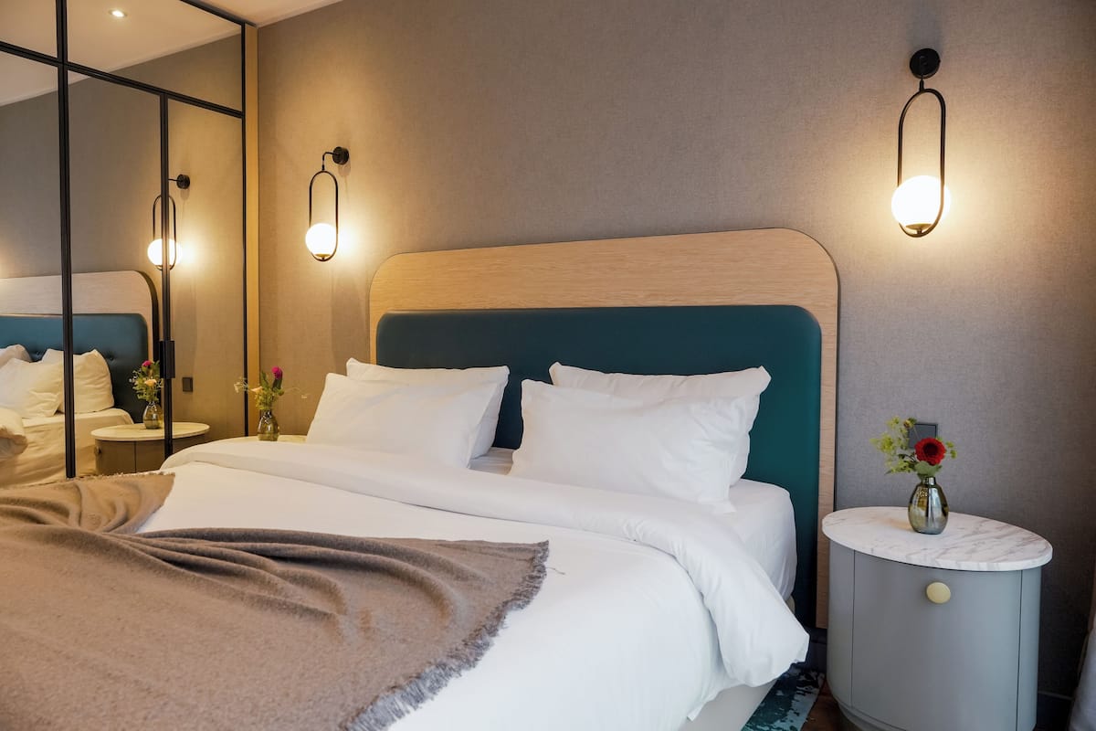 A neatly made bed with white linens and a brown throw blanket, a bedside table with a vase of flowers, and a wall-mounted light fixture.