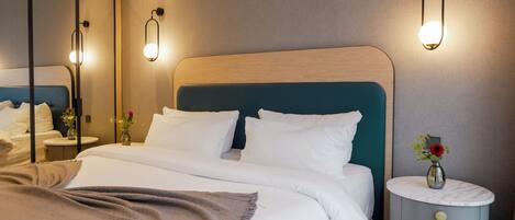 A neatly made bed with white linens and a brown throw blanket, a bedside table with a vase of flowers, and a wall-mounted light fixture.