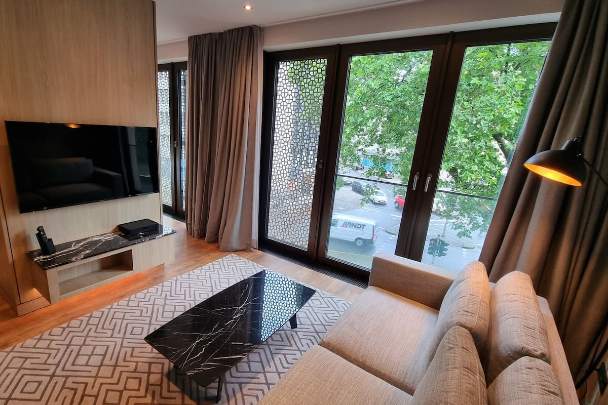 A modern living room with a flat-screen TV, a sofa, a coffee table, and a view of the street through large windows.