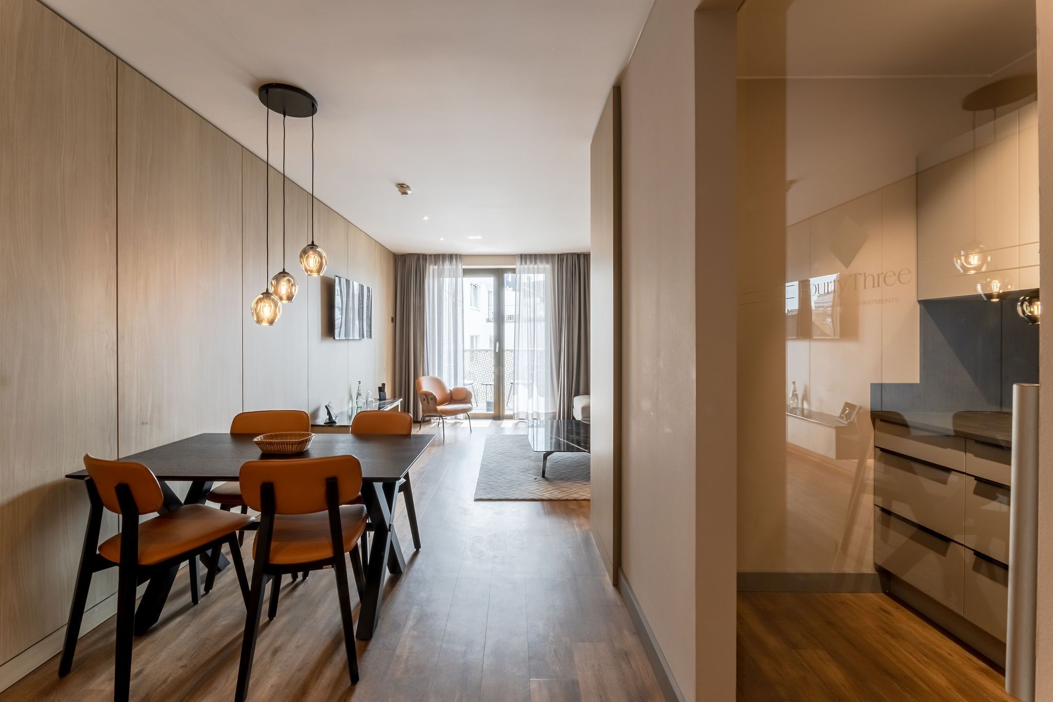 A modern dining area with a wooden table and chairs, a hanging light fixture, and a view of a cityscape through large windows.